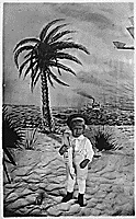 H0016-4 and H0020-2. Gerald R. Ford, Jr. holding a toy boat, poses for portrait; wearing sailor cap, standing on sand, painted backdrop of palm tree, steamship, and airplane in flight, in background. 1916. 