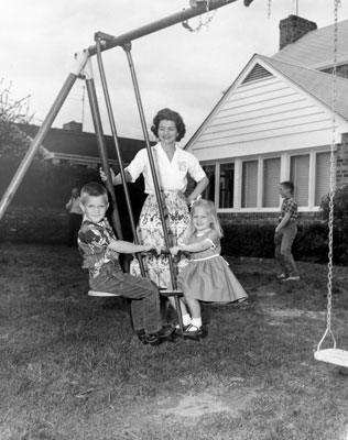 Betty Ford watches as her children Steve and Susan ride on a glider in the back yard of their home. Her eldest son Michael appears in the background.