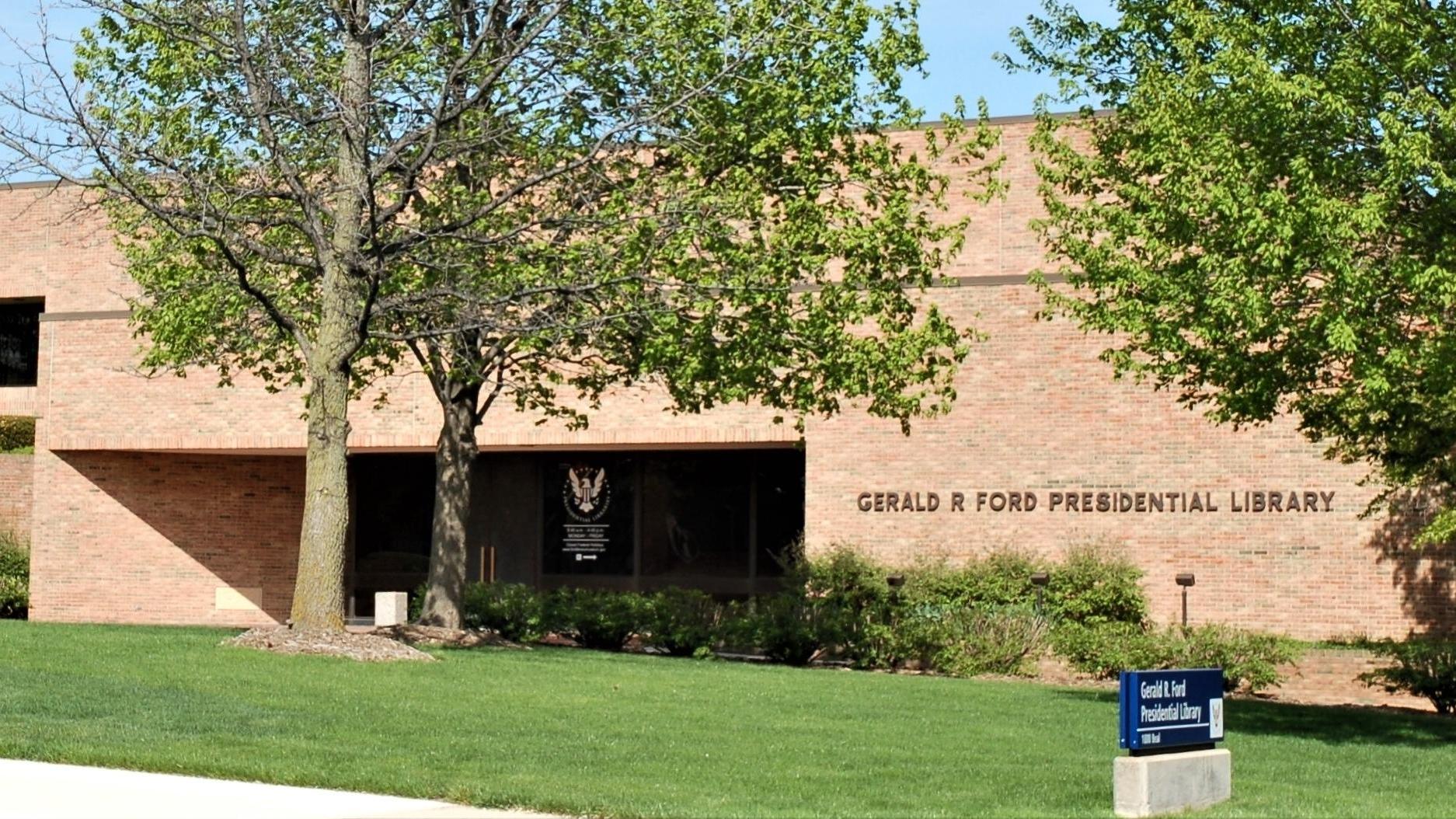The pale brick exterior of the library is shown with an American flag flying to the right of the building and several large trees in front of the building.