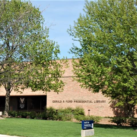 The pale brick exterior of the library is shown with an American flag flying to the right of the building and several large trees in front of the building.