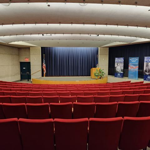 An auditorium with red chairs facing a stage is shown