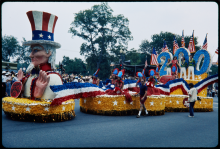 A large, colorful parade float with an Uncle Sam bust and giant 200 drives down a road.