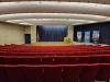 An auditorium with red chairs facing a stage is shown