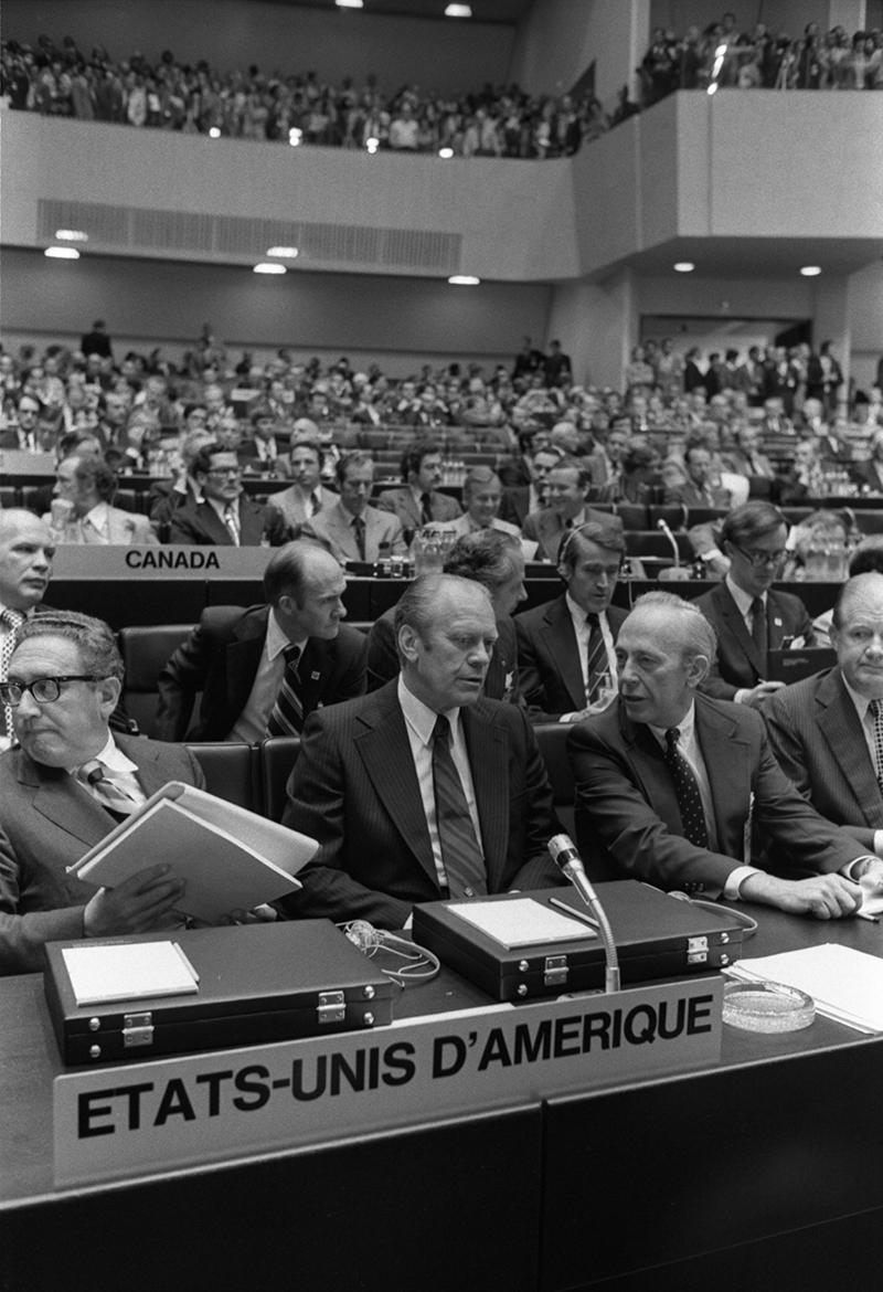 Gerald R. Ford, Kissinger, Hartman are seated at a desk Opening of the Conference on Security and Cooperation in Europe