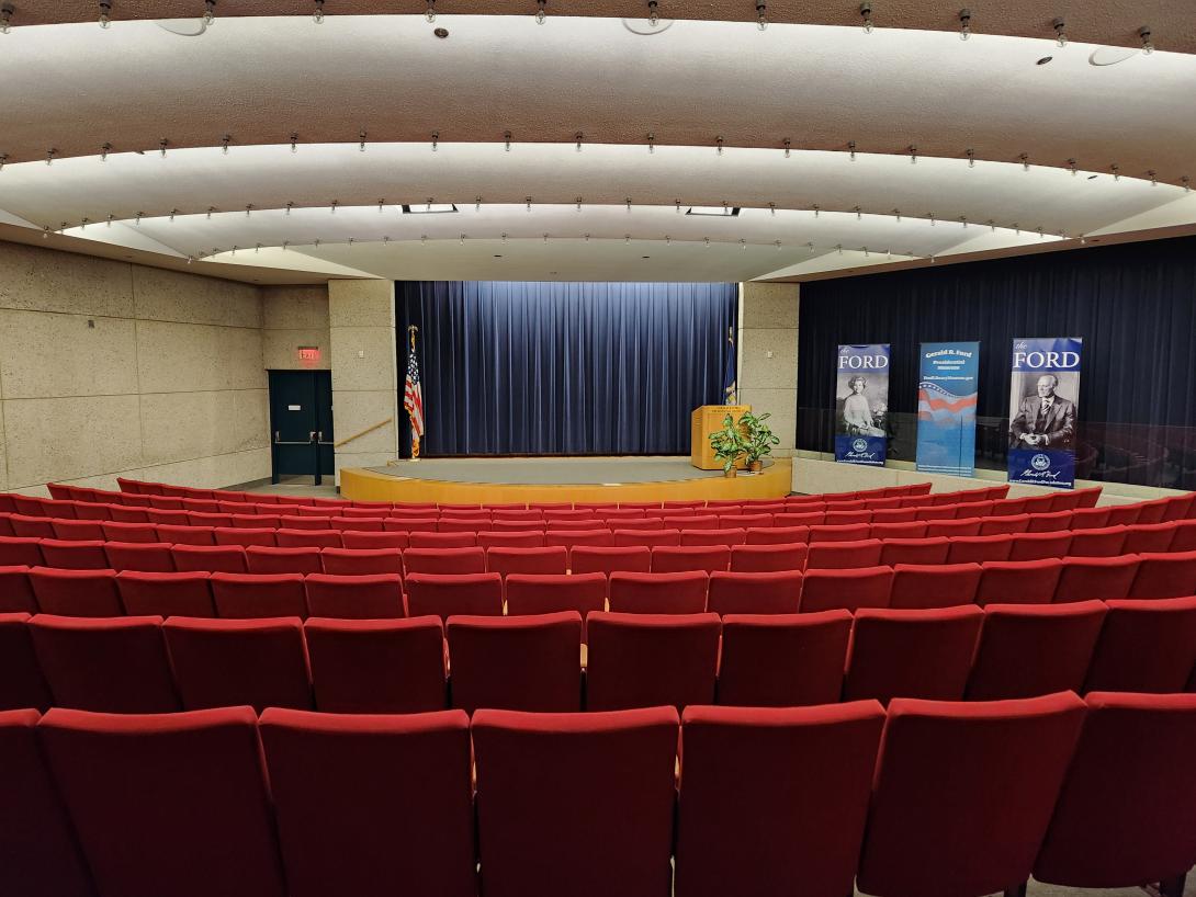 An auditorium with red chairs facing a stage is shown