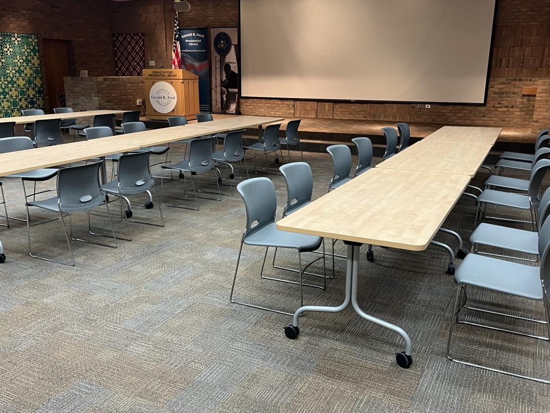 Long rectangular tables with chairs around them are laid out before a stage. 