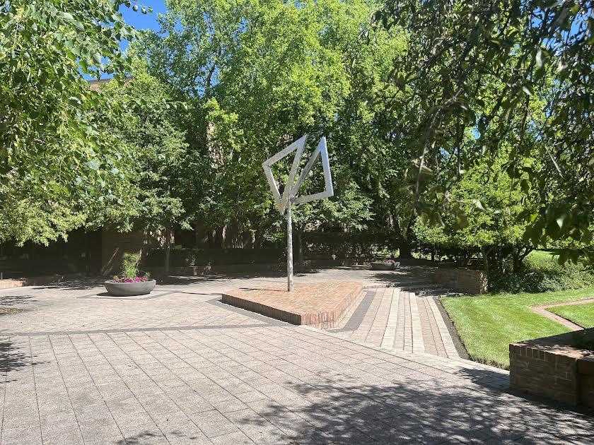 A brick patio with a green grass lawn is shown. A large, silver piece of art stands in the middle.