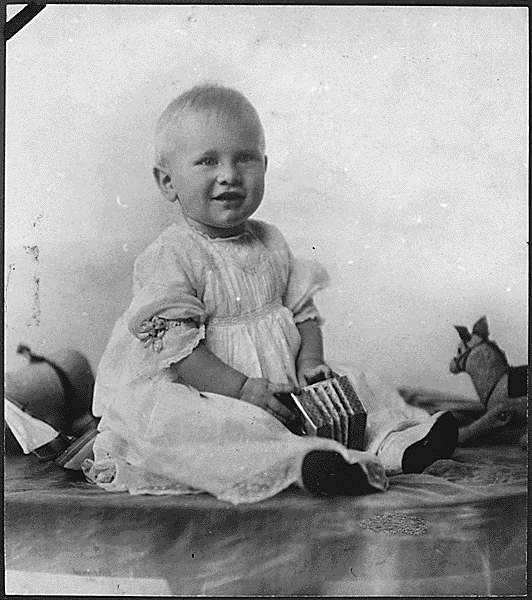 A young baby sits in a white dress, holding a small, toy accordion. 